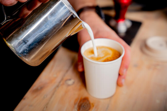 A barista pouring steamed milk into a white paper cup to create latte art.