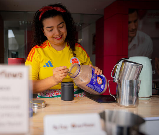 A female barista wearing a yellow Colombia soccer jersey measuring chocolate powder into a cup.