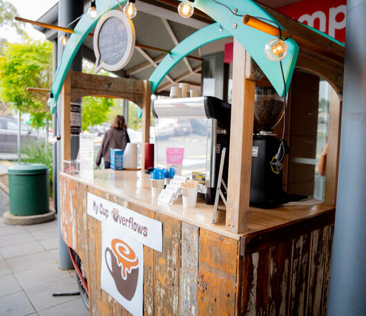 Our custom-built cart fits seamlessly into outdoor markets and private venues across South Australia. A rustic wooden coffee cart with a teal roof set up on an outdoor paved walkway.