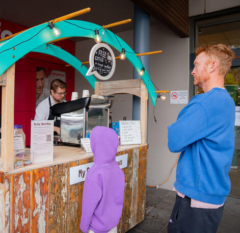A man in a blue sweater and a child waiting at a wooden coffee cart.