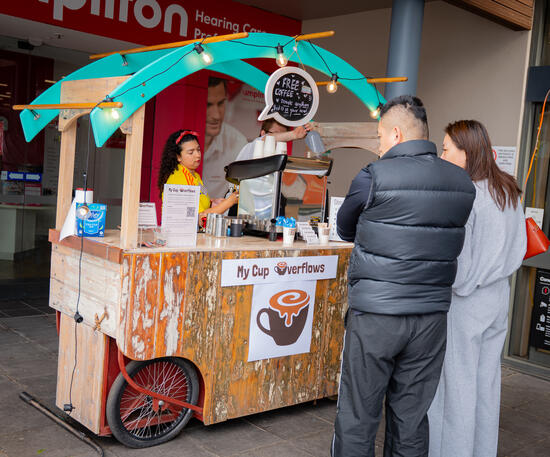 A wooden mobile coffee cart with a teal canopy serving two customers outdoors.