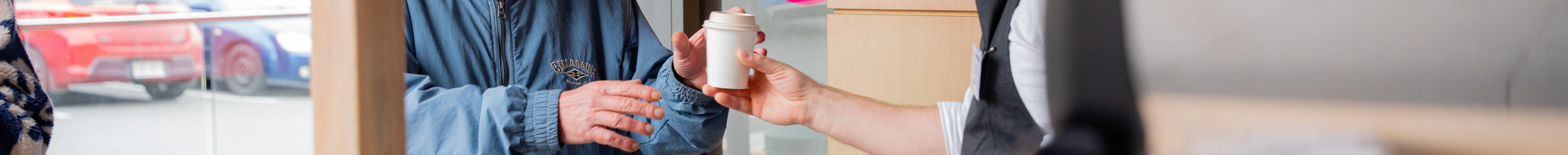 A barista's hand passing a finished white cup of coffee to a customer in a blue jacket.